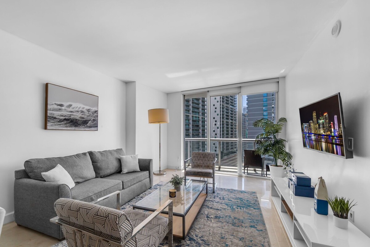 A modern living room with a grey sofa, patterned armchair, glass coffee table, and a large window with a city view.