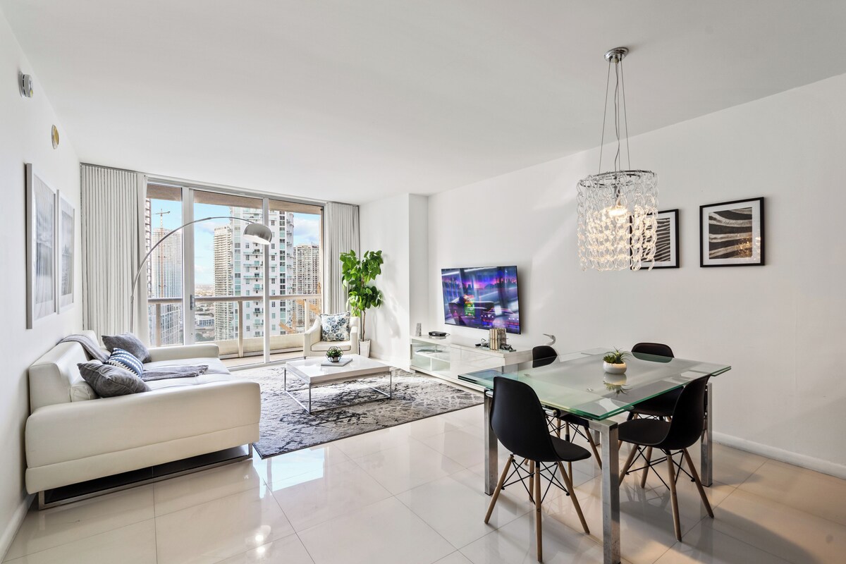 A modern living room with a glass dining table, white sofa, and a large window with a city view.