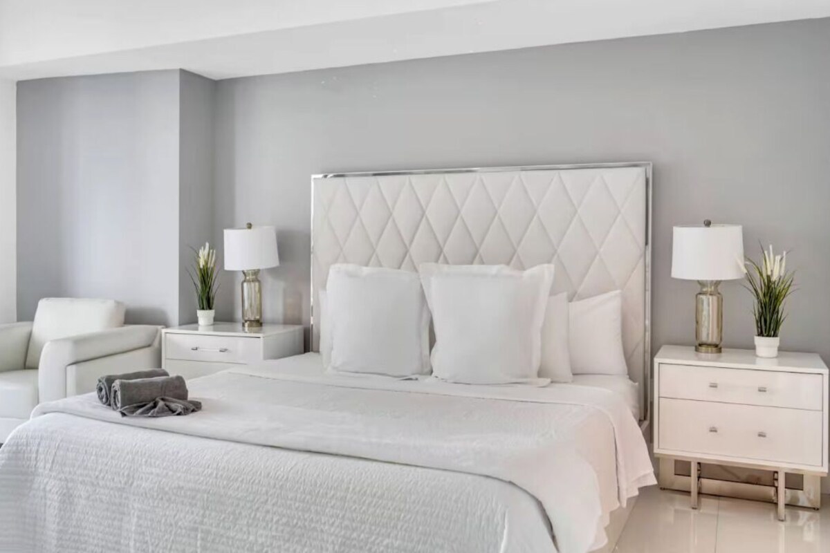 Une chambre à coucher moderne avec un grand lit, deux tables de chevet, une commode, une chaise et une lampe.