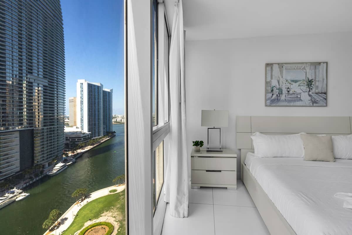 Una habitación de hotel moderna con una cama grande, una mesita de noche, un cuadro y vistas a un río y edificios.