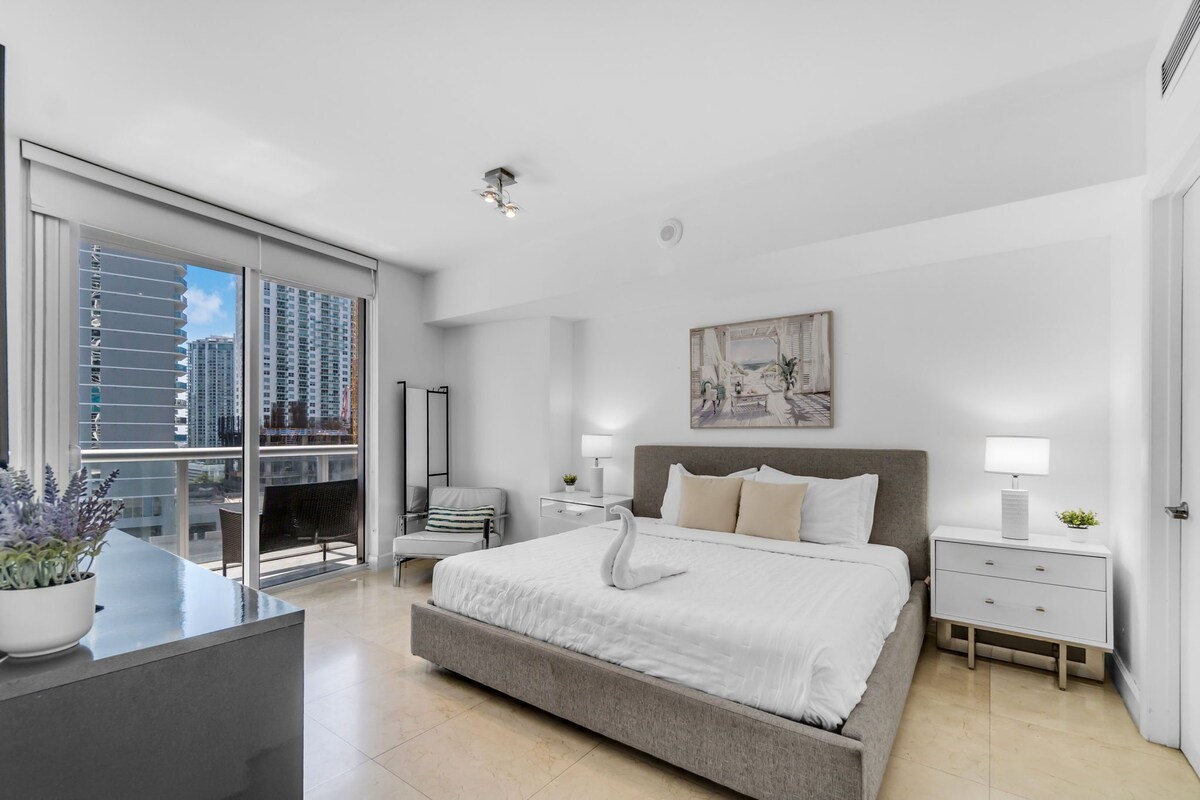 Une chambre moderne avec un grand lit, des tables de chevet, un fauteuil et une vue sur des immeubles de grande hauteur.