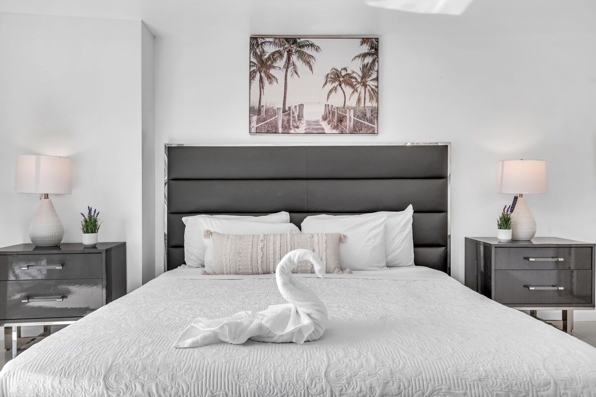 Une chambre moderne avec un grand lit, deux tables de chevet, une tête de lit et une œuvre d’art représentant une scène de plage, accrochée au mur.