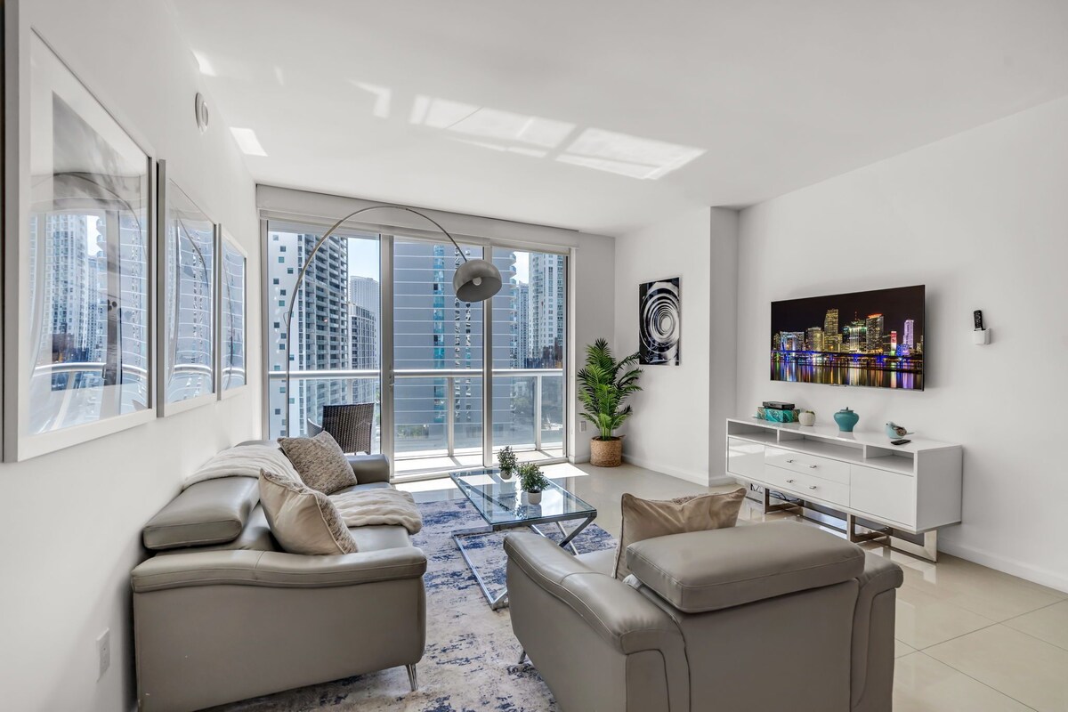A modern living room with a sofa set, a coffee table, a TV mounted on the wall, and a large window with a view of high-rise buildings.