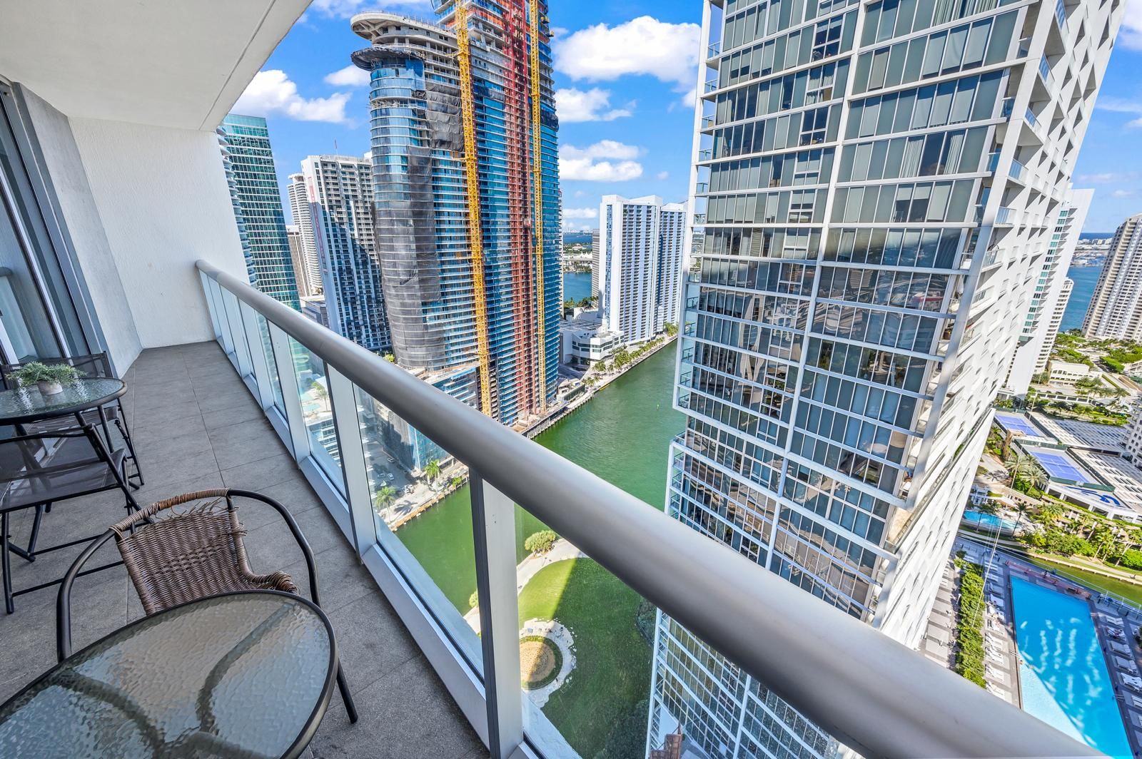 A balcony with a glass table and chair, overlooking a cityscape with modern buildings and a river.