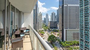 Un balcón con mobiliario de mimbre, con vista a un paisaje urbano con edificios altos y cielo despejado.