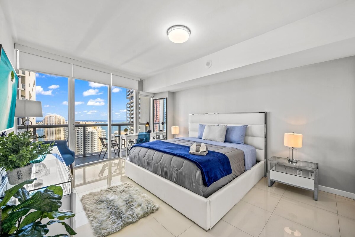 A modern bedroom with a large bed, a desk, and a view of the cityscape.