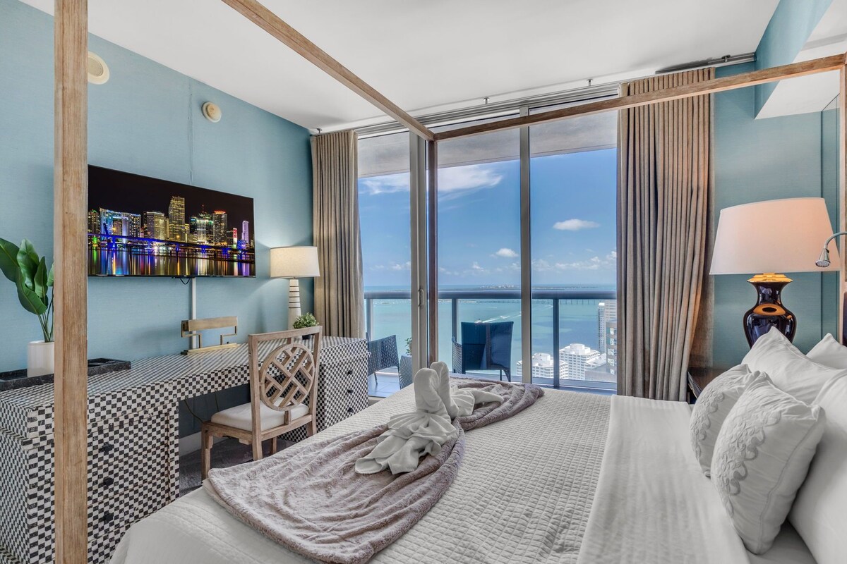 Une chambre d’hôtel avec un grand lit, un bureau avec une chaise, un téléviseur fixé au mur et une vue sur la ville et la mer.