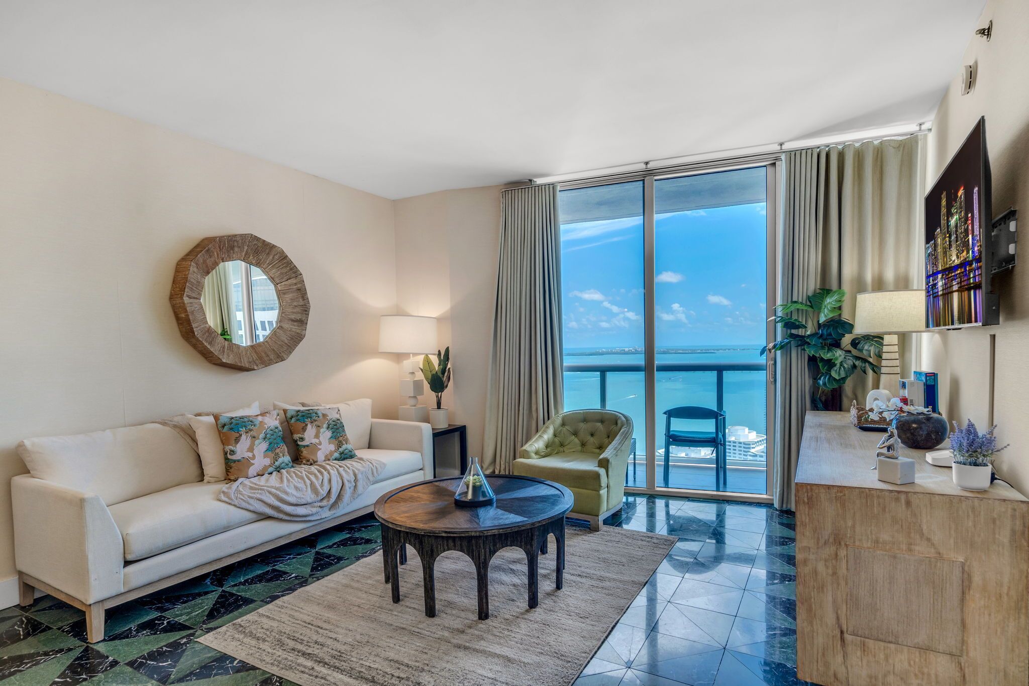 A modern living room with a sofa, a round coffee table, a chair, a lamp, and a view of the ocean through large windows.