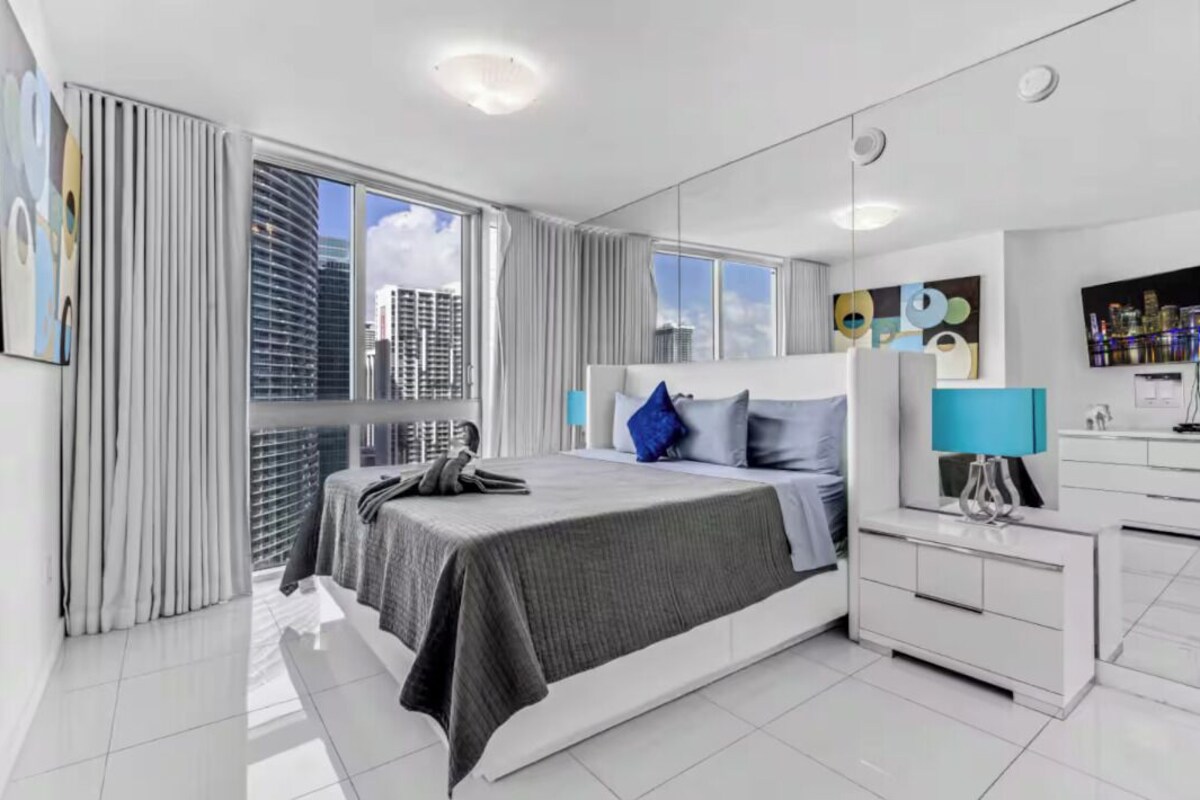 Une chambre d’hôtel moderne dotée d’un grand lit, d’une table de chevet, d’un miroir et offrant une vue sur le paysage urbain grâce à de grandes fenêtres.