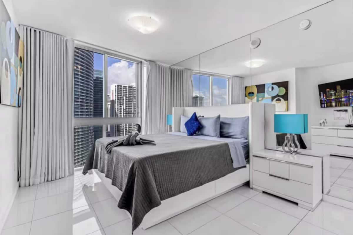 Una habitación de hotel moderna con una cama grande, una mesita de noche, un espejo y vistas de la ciudad a través de amplias ventanas.