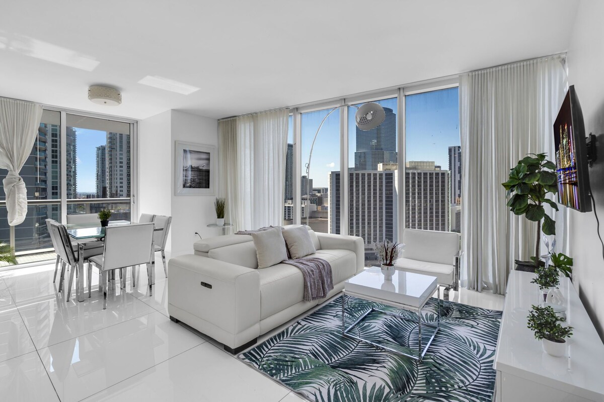 A modern living room with a white sofa, dining table, and chairs, large windows with city views, and a flat-screen TV mounted on the wall.