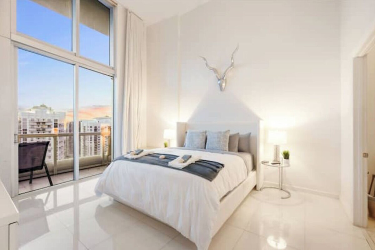Une chambre moderne avec un grand lit, une table de chevet, une décoration murale représentant une tête de cerf et une vue sur les bâtiments à travers de grandes fenêtres.