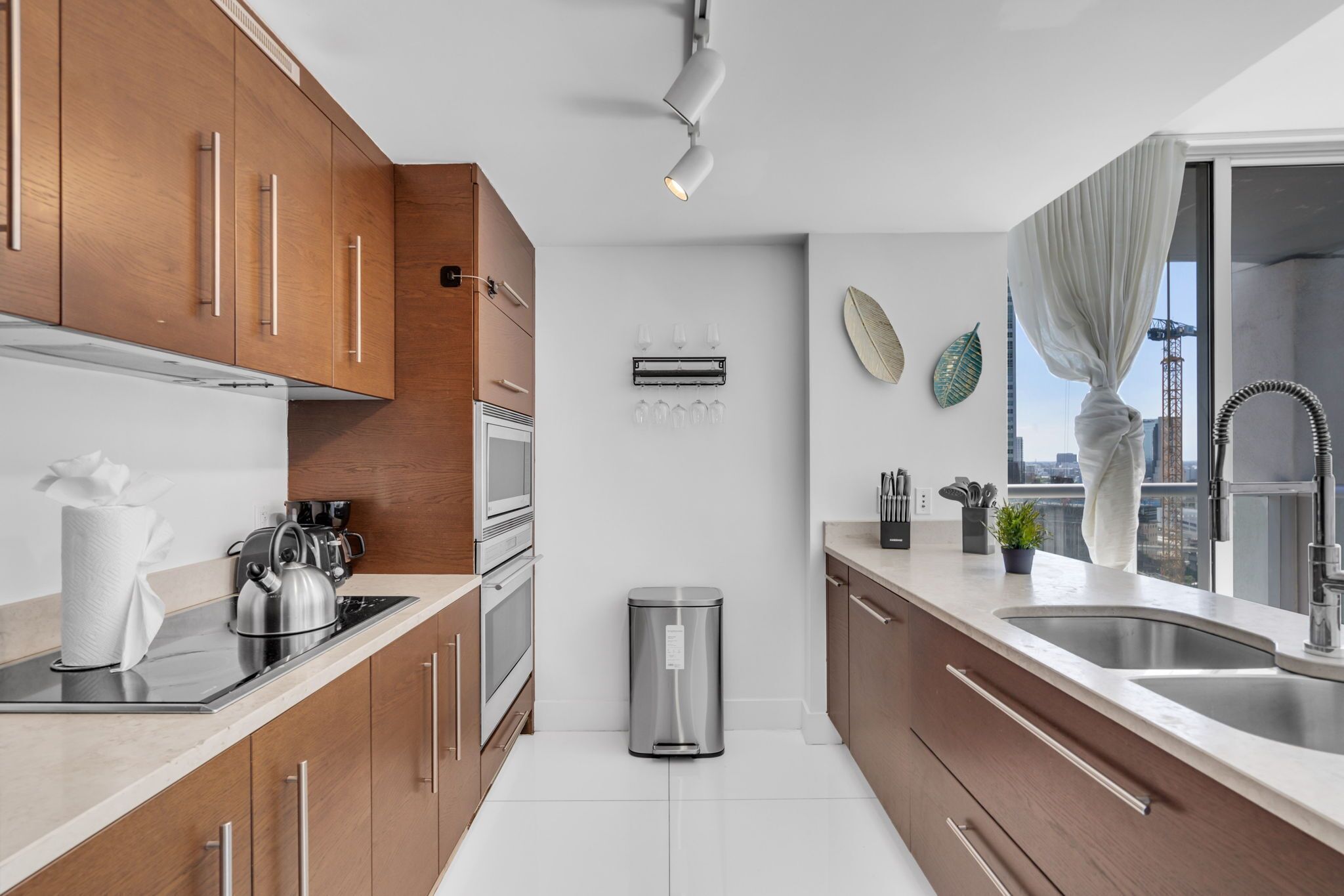 A modern kitchen with wooden cabinets, stainless steel appliances, a trash bin, and a view of the city through large windows.
