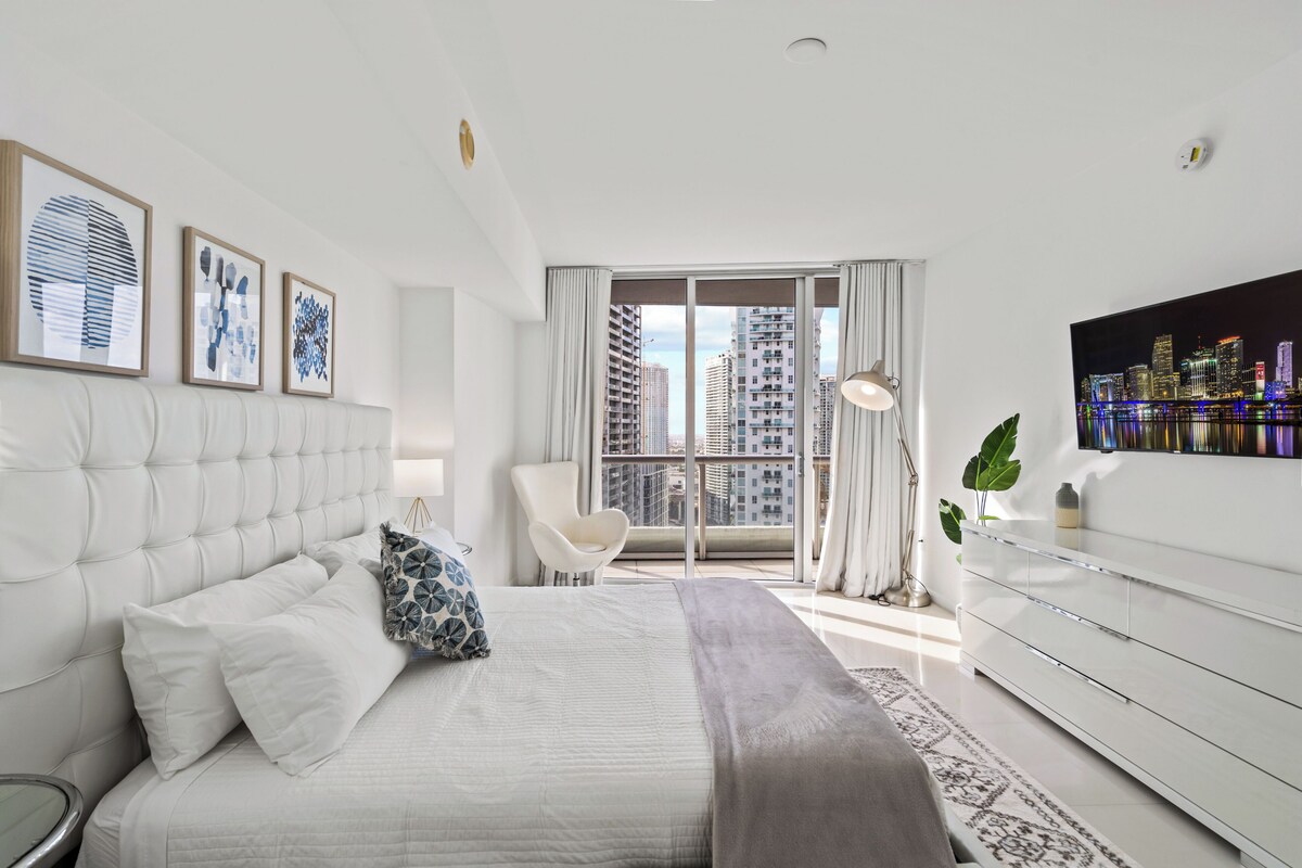 Une chambre moderne avec un lit, une chaise, une commode et une grande fenêtre donnant sur la ville.