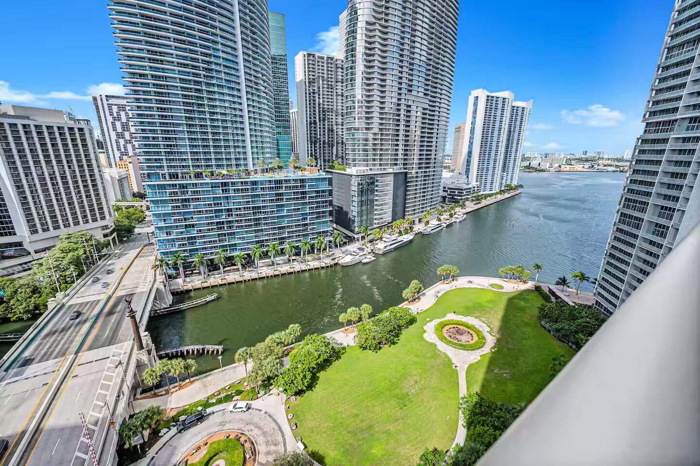 A high-rise urban area with modern buildings, a river, and a park with a circular lawn.