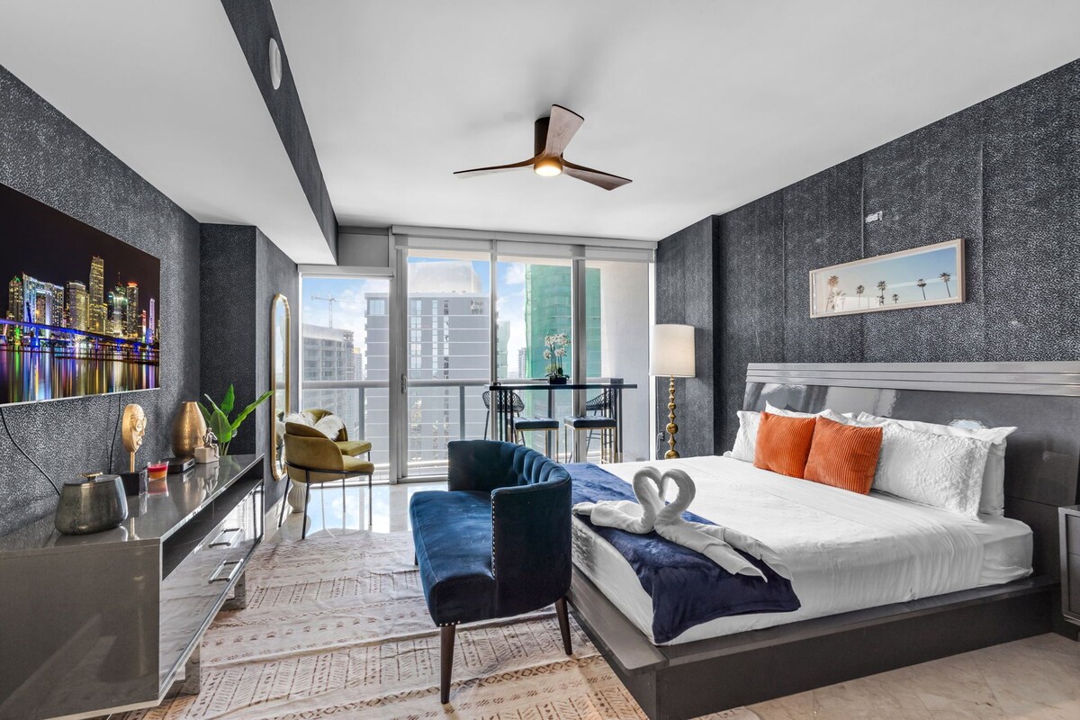 Une chambre d’hôtel moderne avec vue sur la ville, un lit agrémenté d’oreillers décoratifs, un bureau avec une chaise et un balcon avec une table et des chaises.