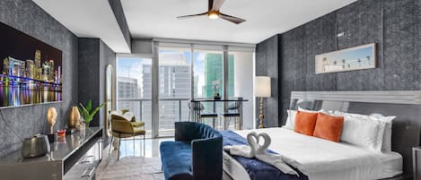A modern hotel room with a cityscape view, a bed with decorative pillows, a desk with a chair, and a balcony with a table and chairs.