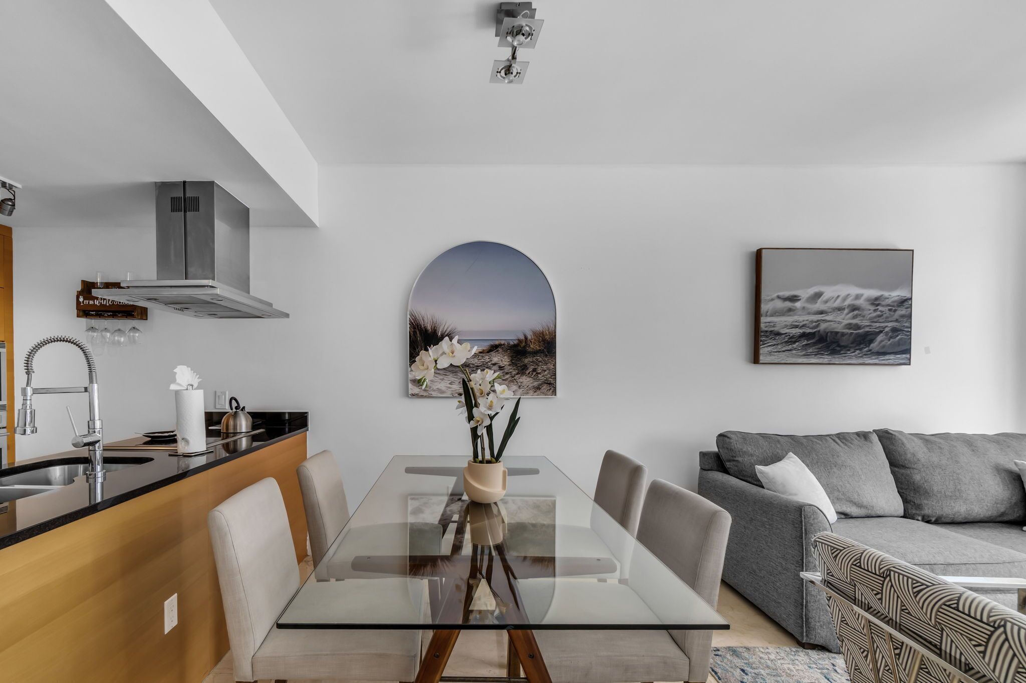 A modern kitchen with a stainless steel range hood, a dining table with chairs, a sofa, and a framed picture on the wall.