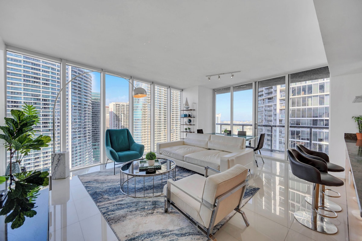 A modern living room with large windows, a white sofa, a teal armchair, and a coffee table.