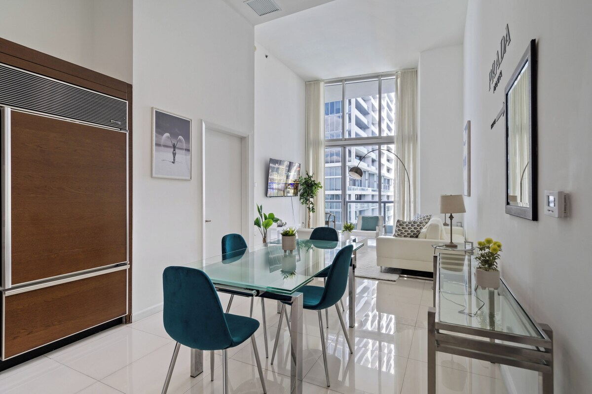 A modern living room with a glass dining table, teal chairs, a white sofa, and a large window with a view of buildings.