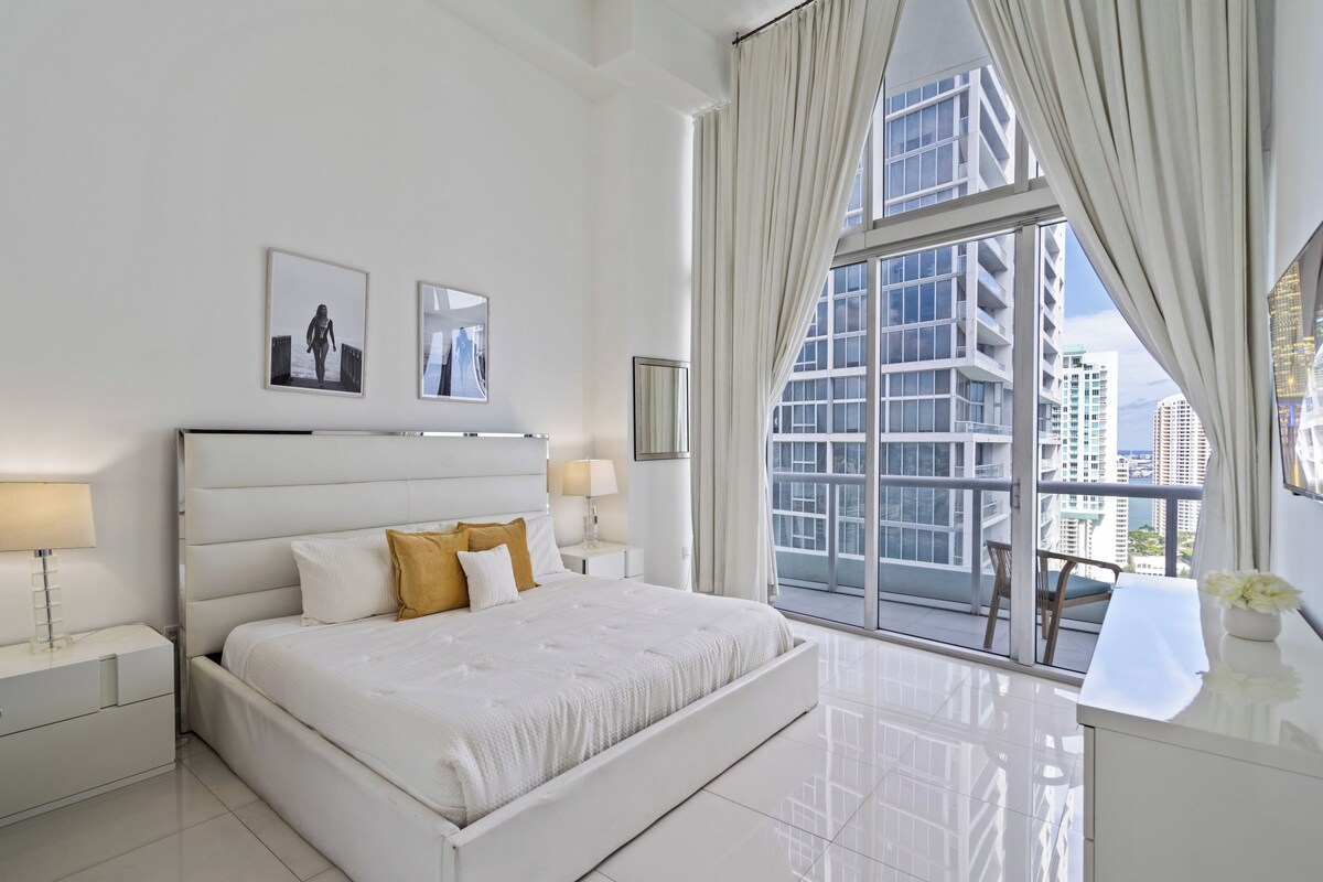 Une chambre d’hôtel moderne, dotée d’un grand lit, de deux tables de chevet, d’une commode, d’une fenêtre avec des rideaux et offrant une vue sur des immeubles de grande hauteur.