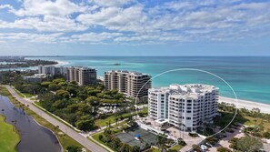 Aerial view of beachfront condos with white facades, curved balconies, tennis courts, and turquoise ocean waters. - The Pierre 906 by FGC (Longboat Key)