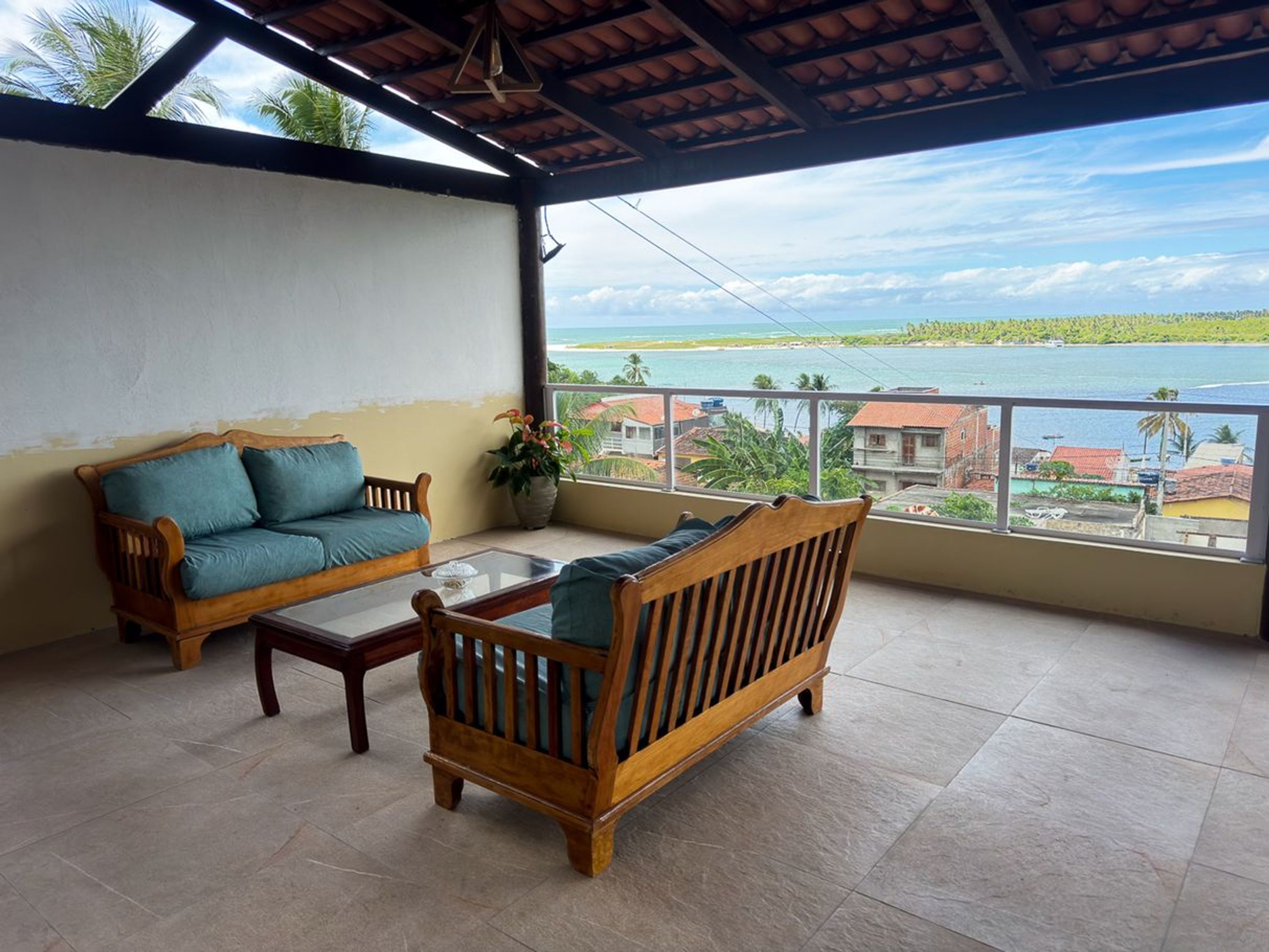 A balcony with a wooden sofa and a small table, overlooking a coastal view.