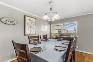 A dining room with a table set for six, wooden chairs, a chandelier, framed artwork, and a window with blinds.