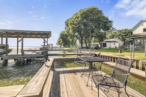 Un muelle de madera con una mesa y sillas, una zona cubierta y un árbol grande al fondo.