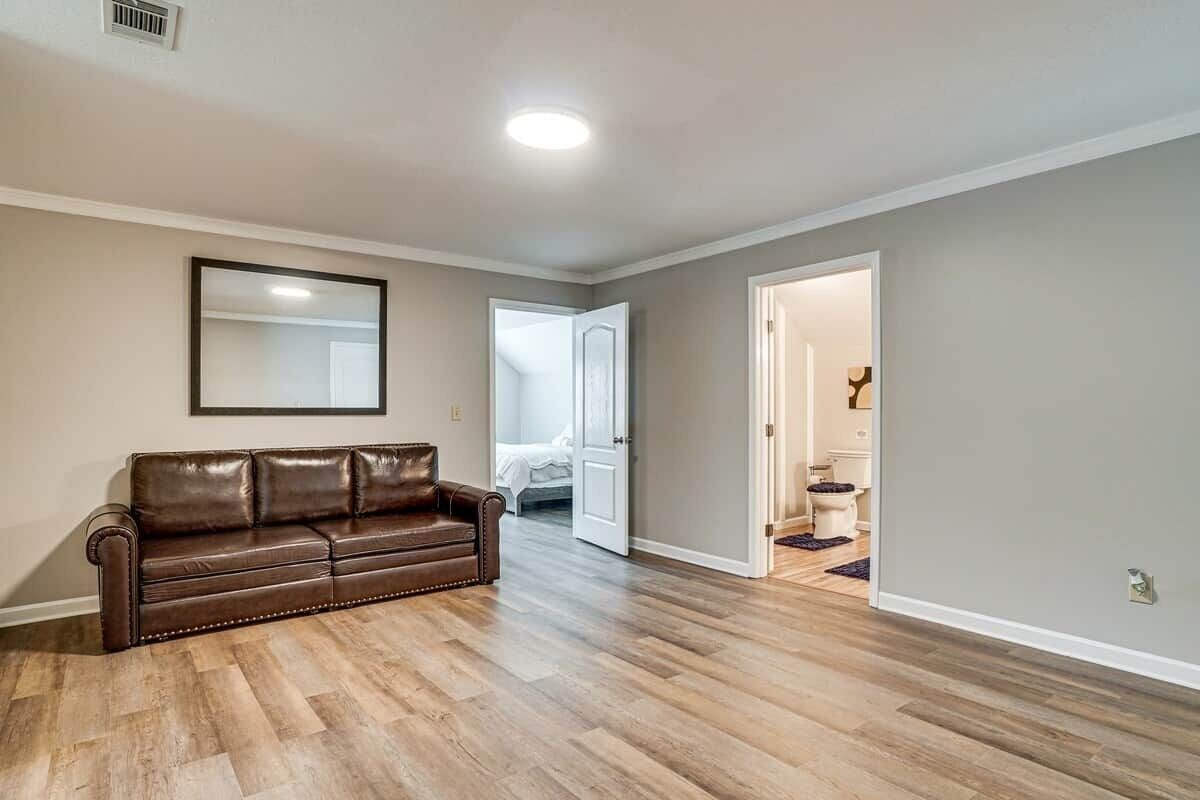 A spacious living room with a brown leather sofa, hardwood flooring, a ceiling light, and a door leading to a bedroom.