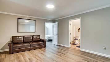 A spacious living room with a brown leather sofa, hardwood flooring, a ceiling light, and a door leading to a bedroom.