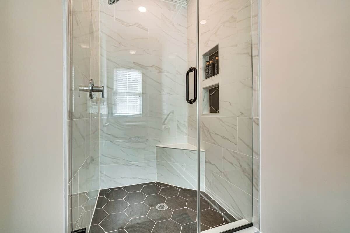A glass-enclosed shower with a marble floor and a white wall with a shelf.