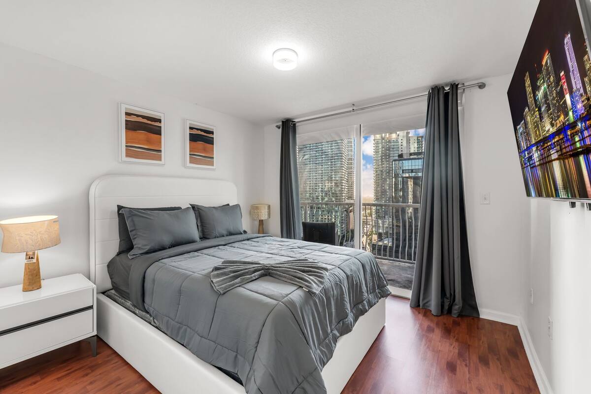 Un dormitorio moderno con cama, mesita de noche y vista a la ciudad por la ventana.