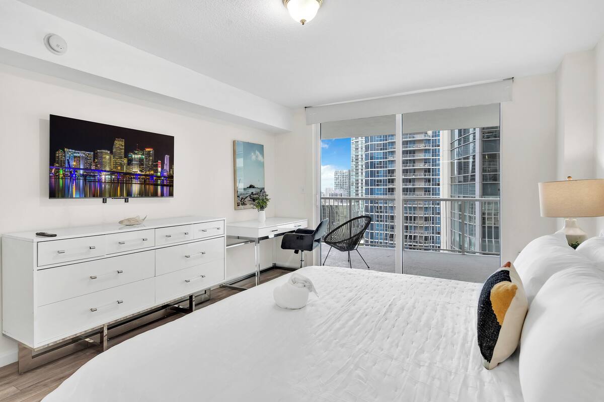 Una habitación de hotel moderna con una cama grande, un escritorio, una silla y un balcón con vistas a la ciudad.