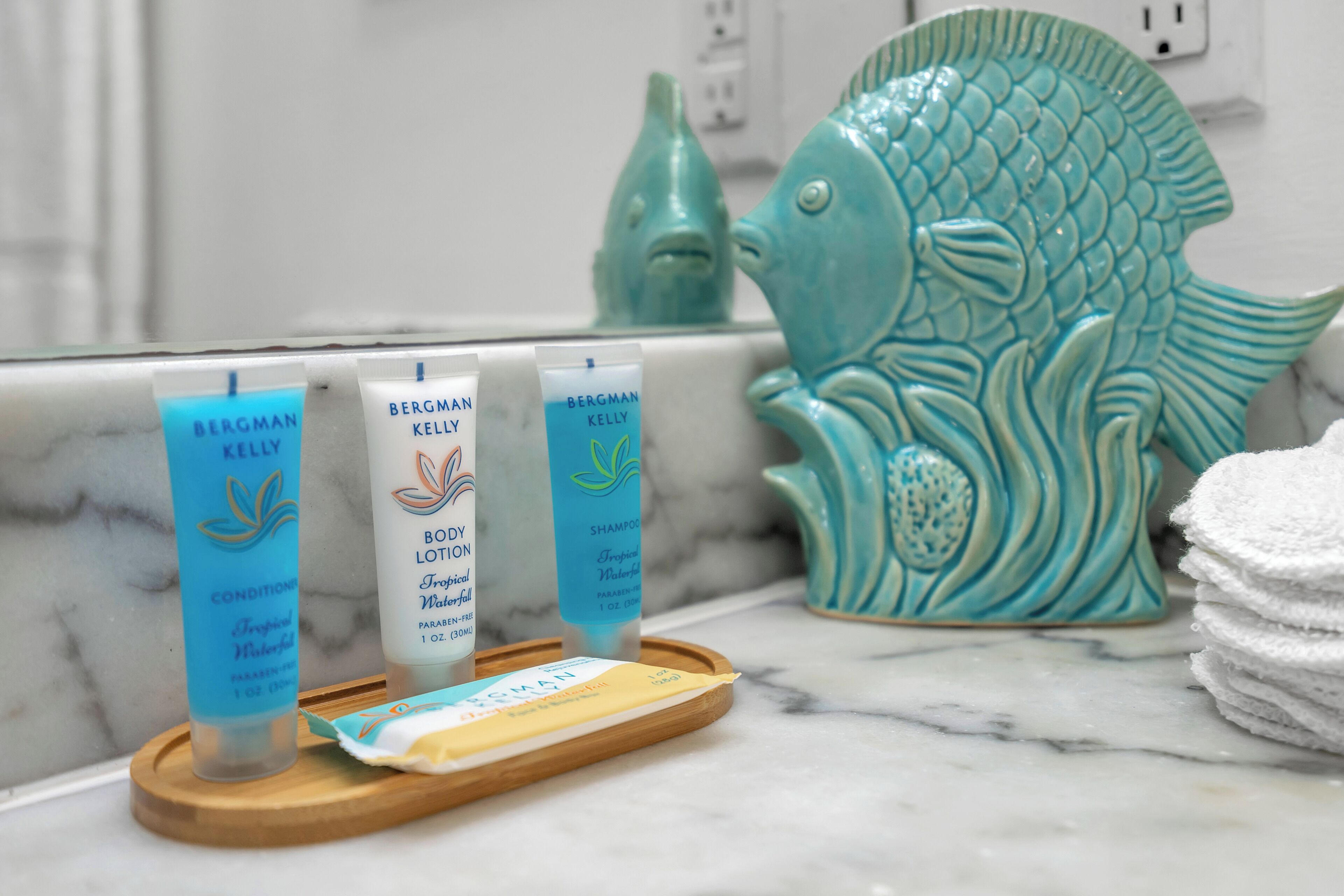 Trois produits de soins de la peau de la marque Bergman Kelly présentés sur un plateau dans une salle de bain, avec une sculpture de poisson décorative.