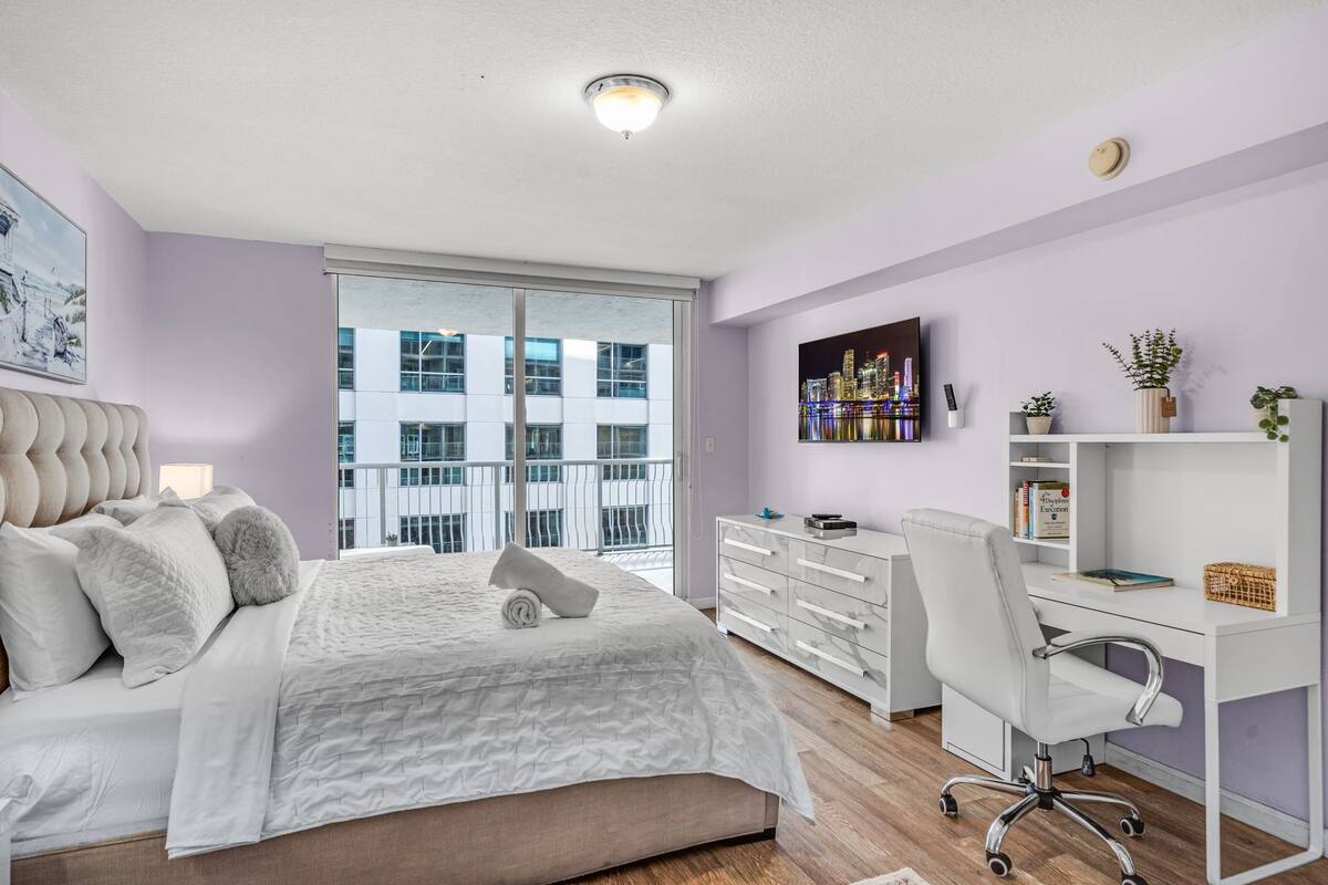 Un dormitorio con cama, escritorio, silla, televisor y ventana con vista a edificios.