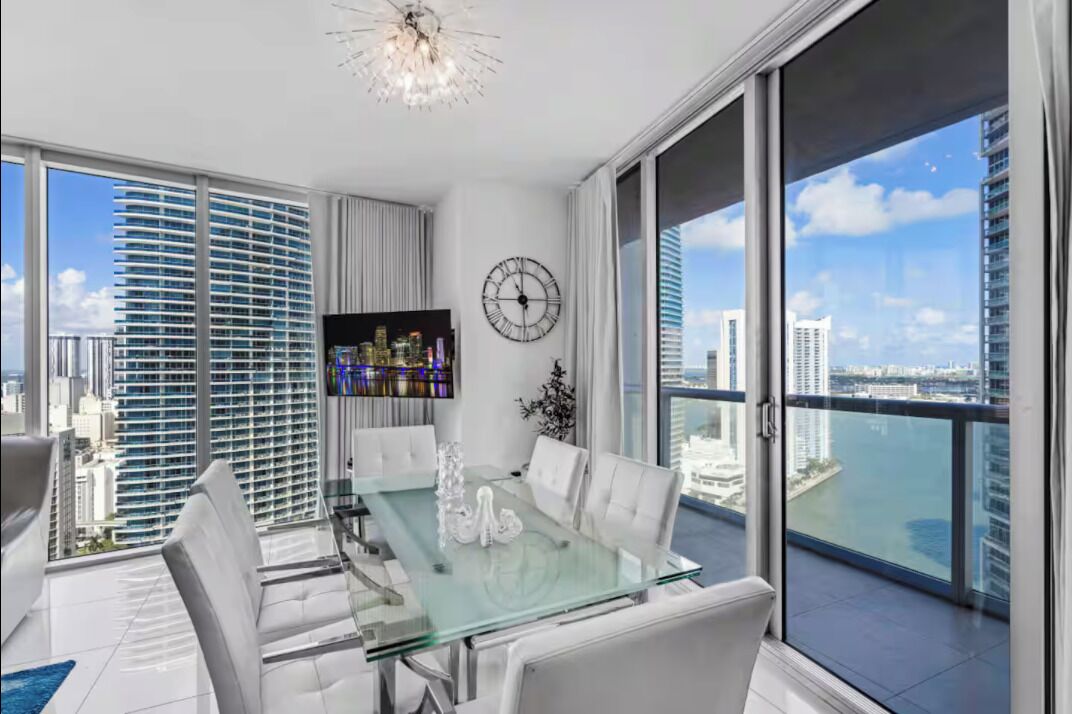 A modern dining area with a glass table, white chairs, a clock, and a view of a cityscape.