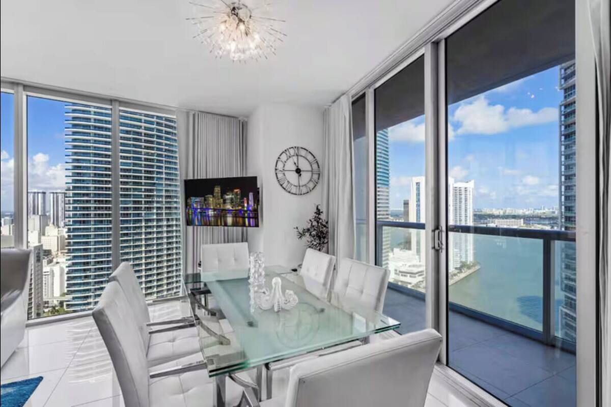 Un comedor moderno con mesa de vidrio, sillas blancas, un reloj y vista a un paisaje urbano.