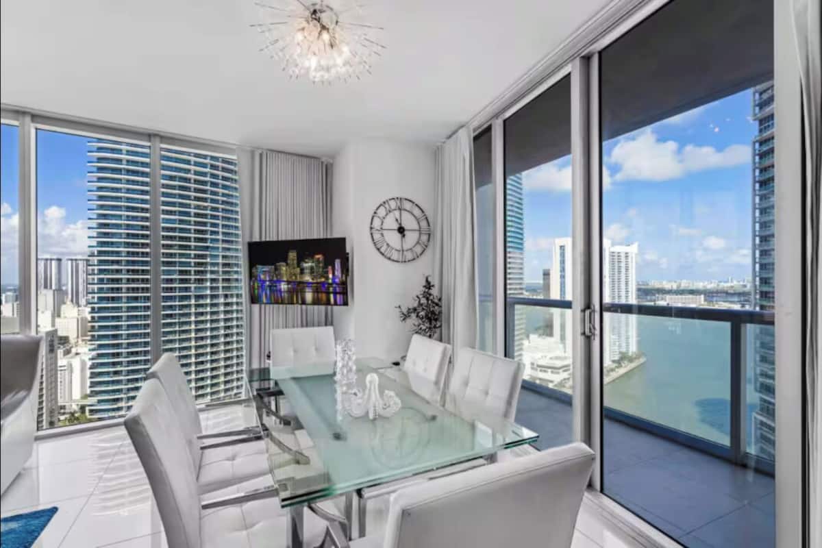 A modern dining area with a glass table, white chairs, a clock, and a view of a cityscape.