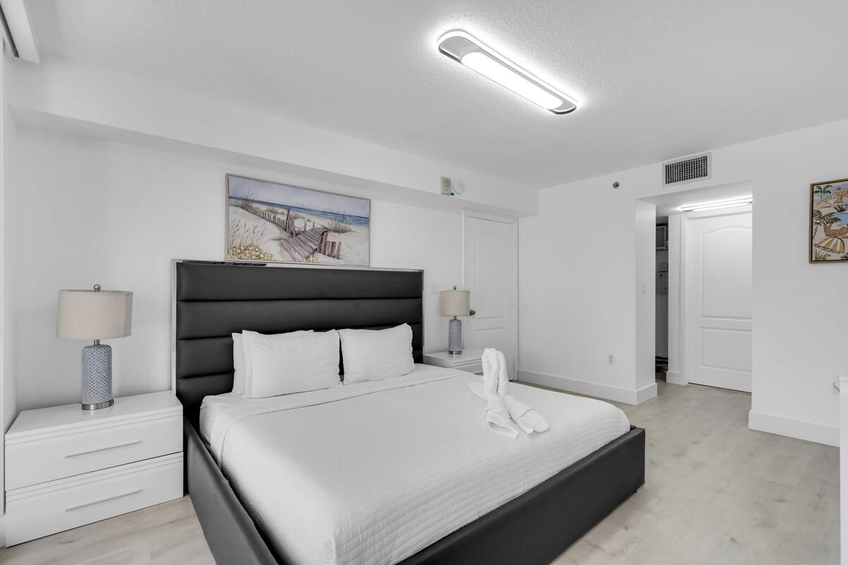 Una habitación de hotel moderna con una cama grande, lámparas de noche, un cuadro en la pared y una puerta que conduce a otra habitación.