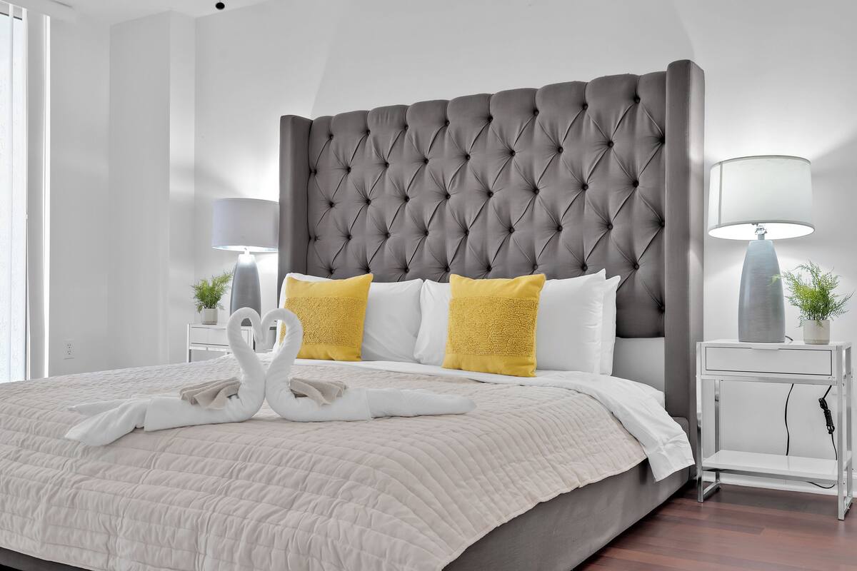 Una cama bien hecha con un cabecero capitonado, almohadas blancas y amarillas, y una toalla dispuesta en forma de cisne.