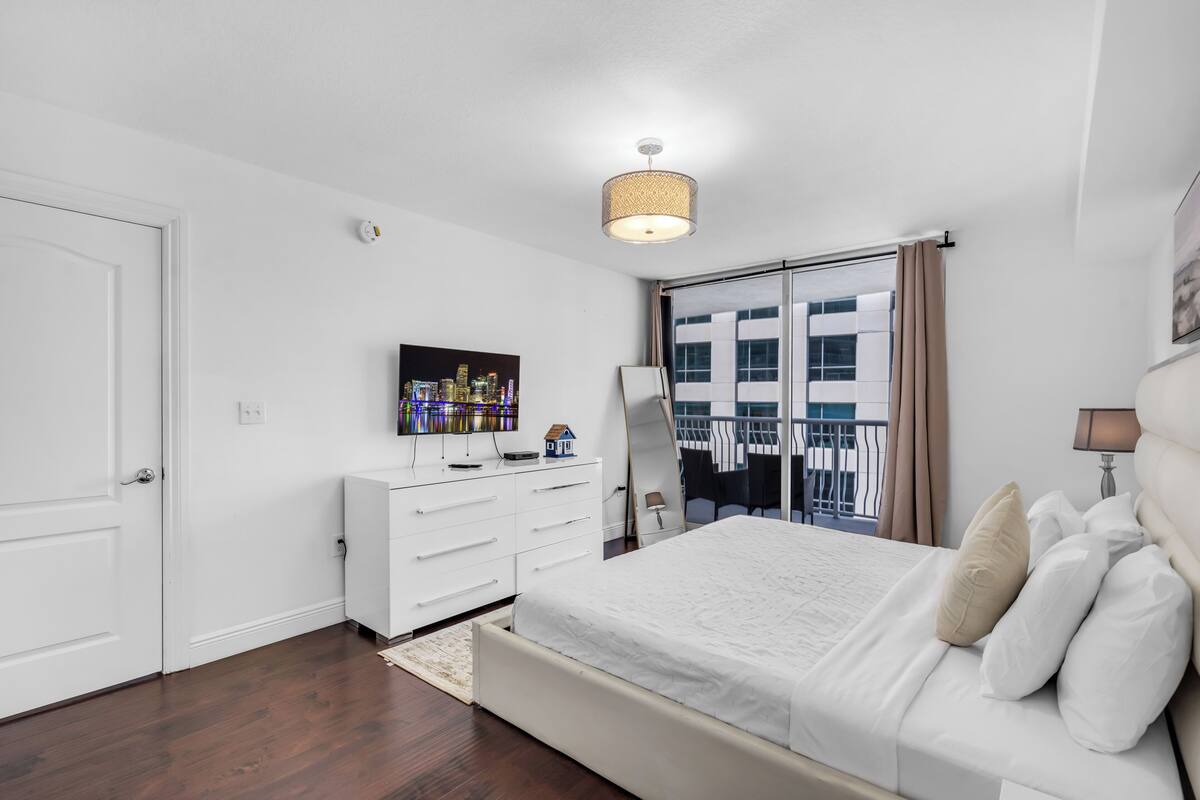 Un dormitorio moderno con una cama grande, un aparador, un televisor de pantalla plana y vistas a la ciudad a través de una puerta corrediza.