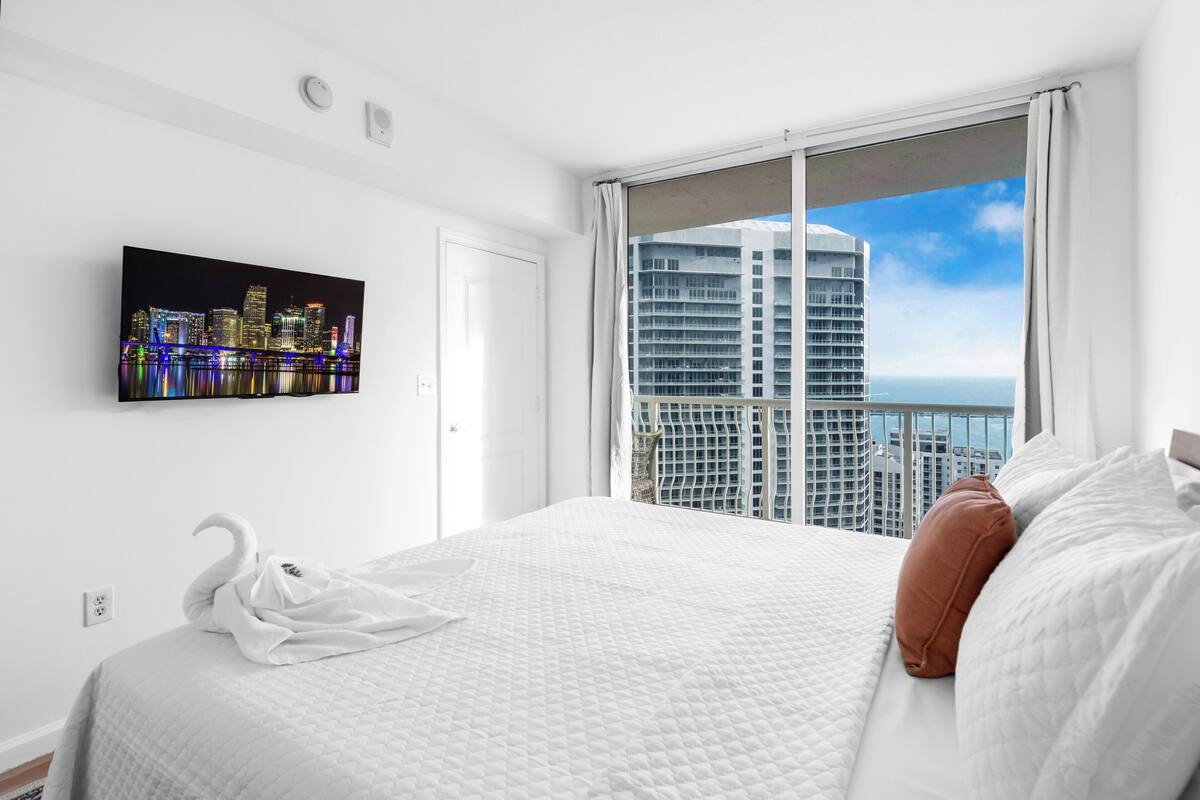 Habitación de hotel con una cama grande, vistas a la ciudad, un televisor de pantalla plana en la pared y una ventana con cortinas.