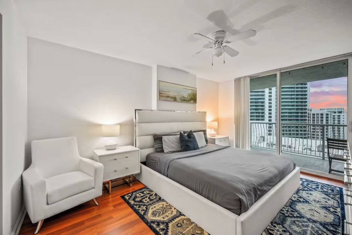 Un dormitorio moderno con una cama grande, un sillón blanco, una mesita de noche con lámpara, un ventilador de techo y un balcón con vista a la ciudad.