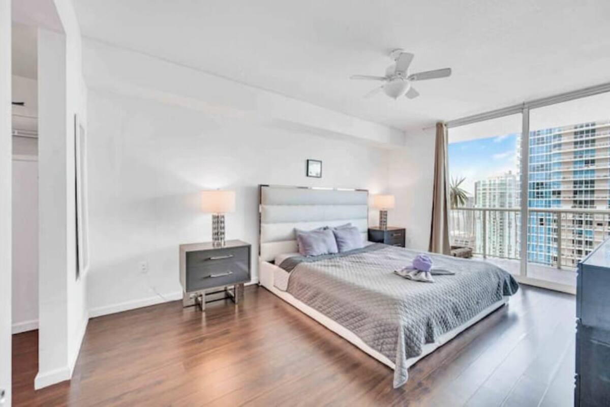 Un dormitorio con cama, mesitas de noche, ventilador de techo y un ventanal con vistas a la ciudad.