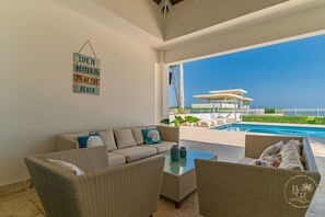 Ein modernes Wohnzimmer mit einer Couch, einem Couchtisch und Korbstühlen mit Blick auf einen Pool und Strand.