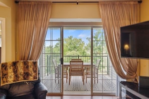 Ein Zimmer mit einer großen Glasschiebetür, die auf eine Außenpatio mit Tisch und Stühlen führt, sowie ein Ledersofa und ein Fernseher auf der rechten Seite.
