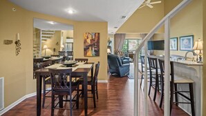 A dining area with a table and chairs, a kitchen in the background, and a view of the outdoors.