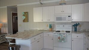 Una cocina con gabinetes blancos, un microondas, un horno y un lavamanos. Hay una pequeña isla con encimera de mármol y un cubo de basura.