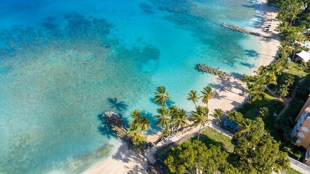 Vista aérea de una playa con aguas turquesas cristalinas, palmeras y un edificio con balcón con vistas a la playa.
